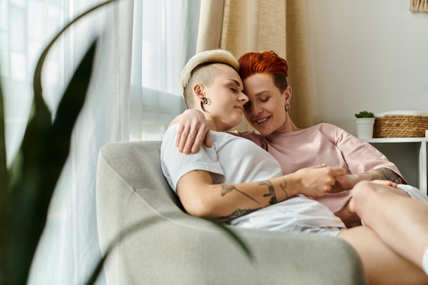 Un momento de corazón entre una pareja lesbiana, sentada en un sofá, abrazándose con amor en un acogedor dormitorio. - Foto, Imagen