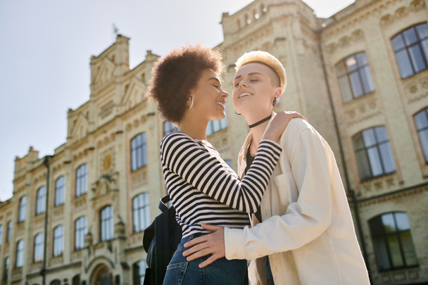 Dwie młode kobiety ubrane stylowo przytulają się przed oszałamiającym budynkiem miejskim, pokazując swoją bliską więź. - Zdjęcie, obraz