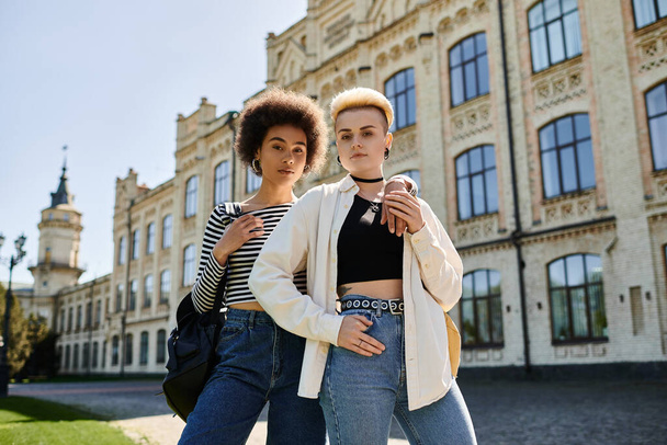 Δύο πολυπολιτισμικές νεαρές γυναίκες με κομψά ρούχα ποζάρουν μπροστά από ένα παλιό κτίριο σε πανεπιστημιούπολη.. - Φωτογραφία, εικόνα