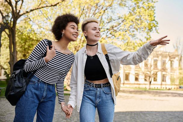 Dos mujeres jóvenes, una pareja lesbiana multicultural, se toman de la mano mientras caminan por una calle cerca de un campus universitario. - Foto, imagen