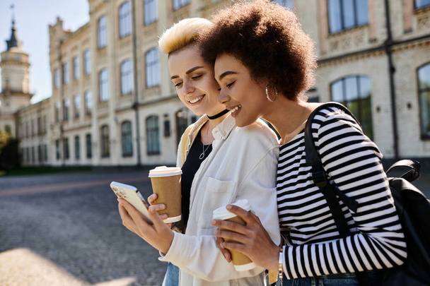 Deux jeunes femmes en tenue élégante, absorbées par un téléphone portable, se tiennent devant un bâtiment moderne dans une rue animée de la ville. - Photo, image