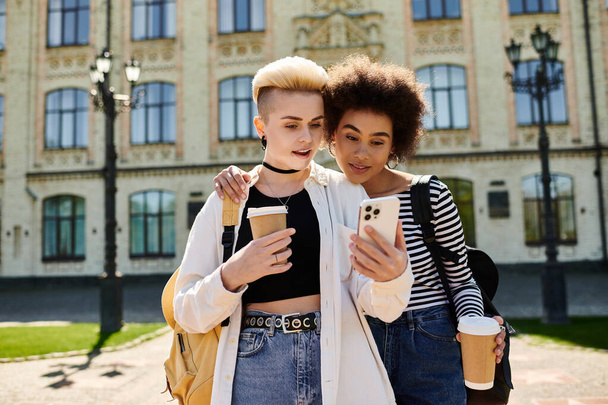 Δύο νεαρές γυναίκες με περιστασιακή ενδυμασία, απορροφημένες από το κινητό τους τηλέφωνο, στέκονται μπροστά από ένα σύγχρονο κτίριο σε ένα πολυσύχναστο δρόμο της πόλης. - Φωτογραφία, εικόνα