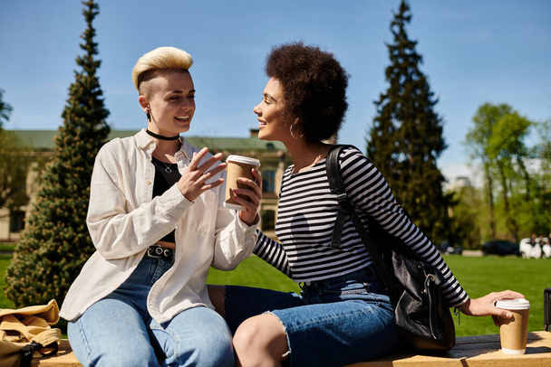 Ένα πολυπολιτισμικό λεσβιακό ζευγάρι, ντυμένο κομψά, κάθεται και συνομιλεί χαρούμενα σε ένα παγκάκι σε ένα πάρκο μια ηλιόλουστη μέρα. - Φωτογραφία, εικόνα