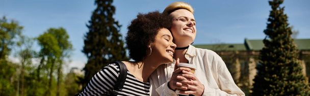 Δύο νεαρές γυναίκες, ένα πολυπολιτισμικό λεσβιακό ζευγάρι, στέκονται σε ένα πάρκο αγκαλιάζοντας η μία την άλλη θερμά. - Φωτογραφία, εικόνα