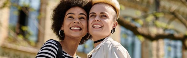 Zwei junge Frauen, ein multikulturelles lesbisches Paar, teilen ein freudiges Lächeln vor einem Universitätsgebäude. - Foto, Bild