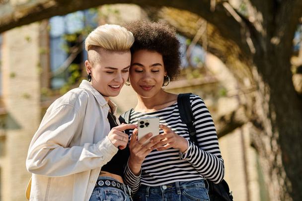 Дві молоді жінки, випадково одягнені, захоплені в мобільний телефон, стоять разом на відкритому повітрі. - Фото, зображення