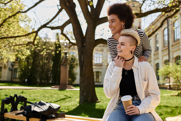 Twee jonge vrouwelijke vrienden in casual kleding zitten op een bankje in het park, genieten van elkaars gezelschap op een zonnige dag. - Foto, afbeelding