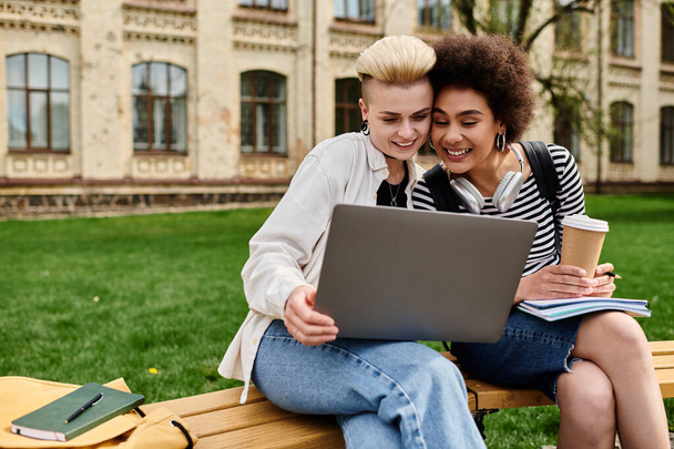 Two women in casual attire sitting on a bench, focused on a laptop screen, in an urban setting. - Photo, Image