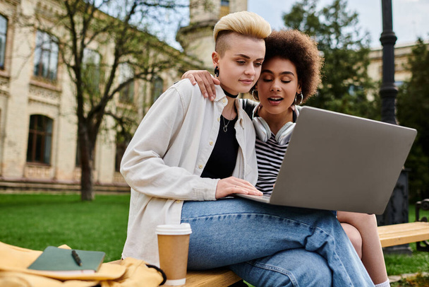 Zwei junge Frauen in lässiger Kleidung sitzen auf einer Bank, in ihren Laptop-Bildschirm vertieft. - Foto, Bild