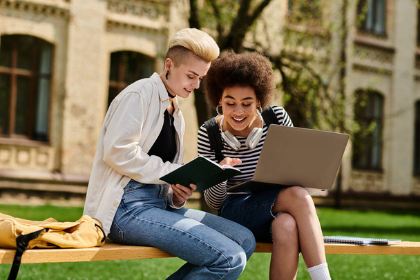 Dwie młode kobiety, stylowo ubrane, pracujące razem na laptopie siedząc na ławce na świeżym powietrzu. - Zdjęcie, obraz