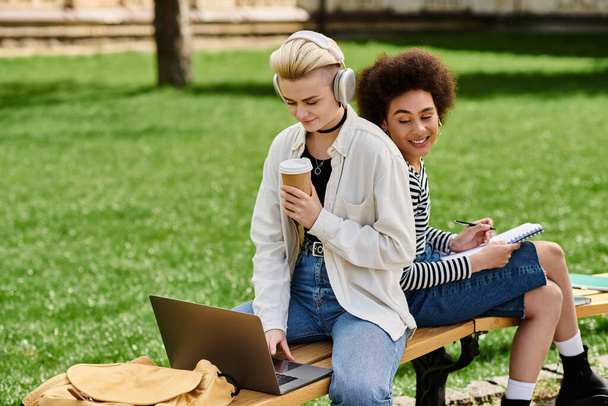 Twee meisjes, terloops gekleed, zitten op een bankje buiten, verzonken in een laptop. - Foto, afbeelding