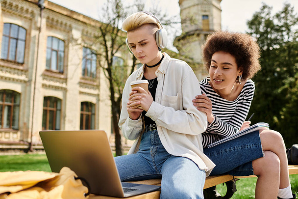 Zwei Frauen in Freizeitkleidung sitzen auf einer Bank, konzentrieren sich auf den Laptop-Bildschirm und arbeiten gemeinsam im Freien. - Foto, Bild