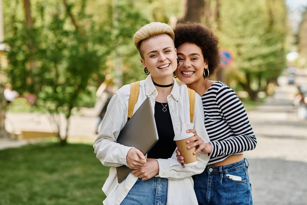 Zwei junge Frauen in Freizeitkleidung stehen in einem Park und halten an einem sonnigen Tag Tassen Kaffee in der Nähe eines Universitätscampus. - Foto, Bild