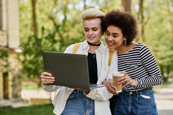 Dwie młode kobiety, przypadkowo ubrane, siedzą obok siebie w parku, skupione na ekranie laptopa, zaangażowane w rozmowę. - Zdjęcie, obraz