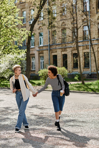 Zwei junge Frauen gehen Händchen haltend eine Kopfsteinpflasterstraße in der Nähe eines Universitätscampus entlang. - Foto, Bild