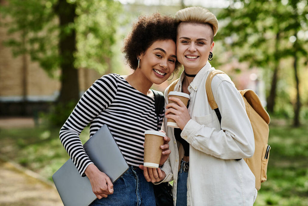 公園の設定でチャットしながらコーヒーカップとラップトップを保持するカジュアルな服装の2人の若い女性. - 写真・画像