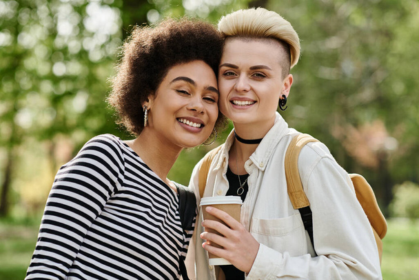 Dos mujeres jóvenes, elegantemente vestidas, felizmente sostienen tazas de café en un vibrante entorno de parque cerca de un campus universitario. - Foto, imagen