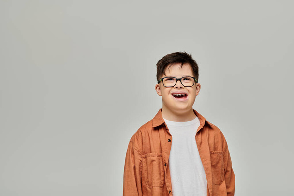 Mały chłopiec z zespołem Downa, noszący okulary, stoi na szarym tle.. - Zdjęcie, obraz