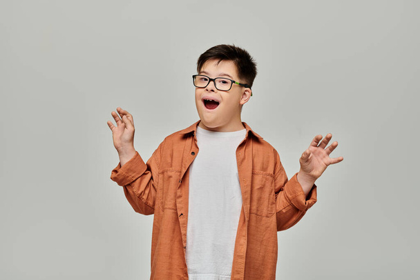 Un niño con síndrome de Down en gafas sostiene sus manos sobre un fondo gris. - Foto, imagen