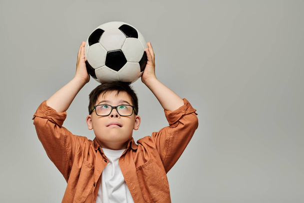 Un charmant petit garçon trisomique tient joyeusement un ballon de foot au-dessus de sa tête. - Photo, image
