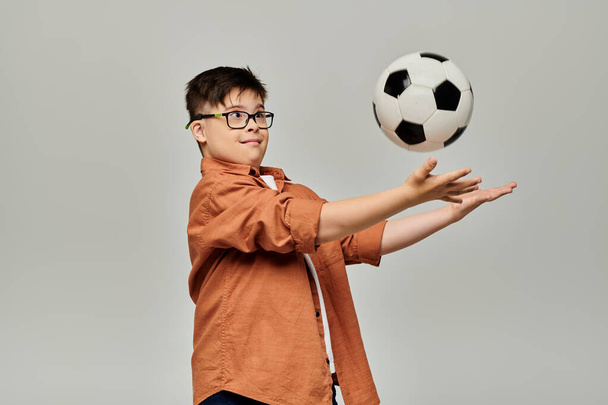 Un niño encantador con síndrome de Down equilibra una pelota de fútbol sobre un fondo gris. - Foto, Imagen