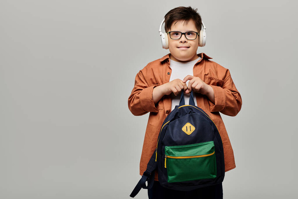 mały chłopiec z zespołem Downa noszący słuchawki, trzymający plecak. - Zdjęcie, obraz