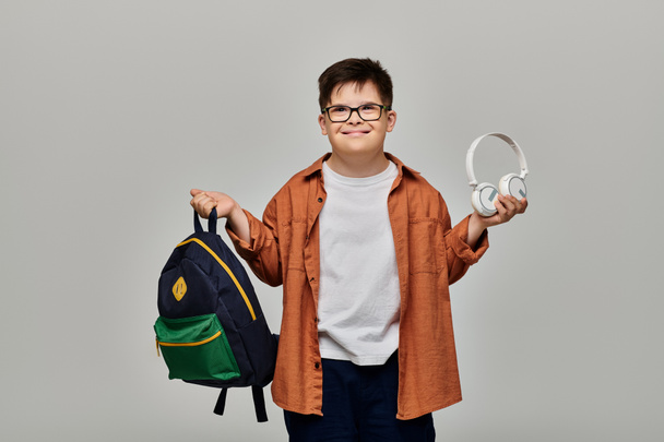 Mały chłopiec z zespołem Downa trzymający plecak i noszący słuchawki. - Zdjęcie, obraz