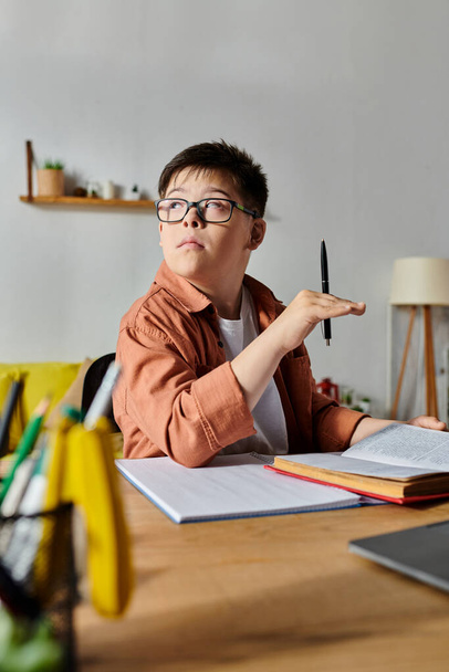 Ein entzückender Junge mit Down-Syndrom sitzt an einem Schreibtisch und hält einen Stift. - Foto, Bild