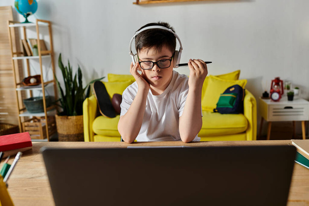 Um menino com síndrome de Down senta-se em uma mesa com fones de ouvido, focando em seu laptop. - Foto, Imagem