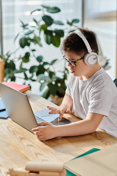 Uroczy chłopiec z zespołem Downa noszący słuchawki jest pochłonięty używaniem laptopa w domu.. - Zdjęcie, obraz