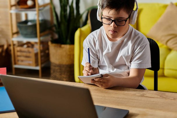 Un adorabile ragazzo con la sindrome di Down è assorto nell'uso di un computer portatile e indossa cuffie a un tavolo. - Foto, immagini