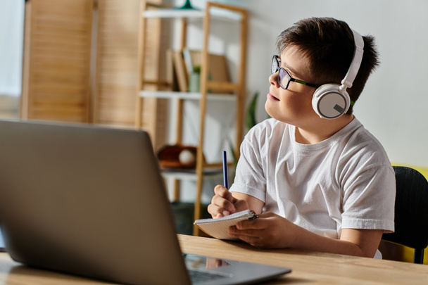Um menino adorável com síndrome de Down usando fones de ouvido se senta em uma mesa usando um laptop. - Foto, Imagem