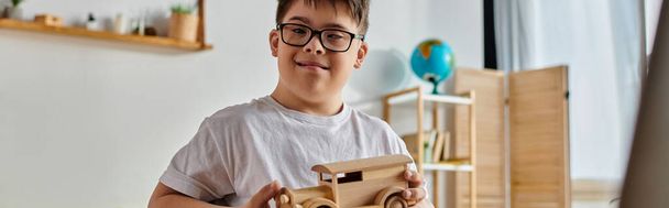 Un niño con síndrome de Down con gafas juega con un coche de juguete de madera. - Foto, Imagen