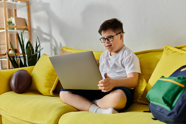 Μικρό αγόρι με σύνδρομο Down χρησιμοποιώντας φορητό υπολογιστή σε κίτρινο καναπέ. - Φωτογραφία, εικόνα