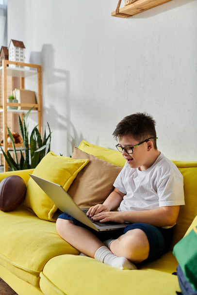 Ένα αγόρι με σύνδρομο Down χρησιμοποιεί ένα φορητό υπολογιστή σε ένα φωτεινό κίτρινο καναπέ. - Φωτογραφία, εικόνα