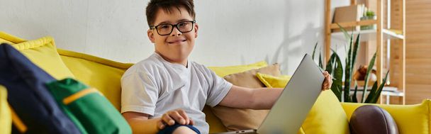 imádnivaló fiú Down-szindróma ül a sárga kanapén, a laptop. - Fotó, kép