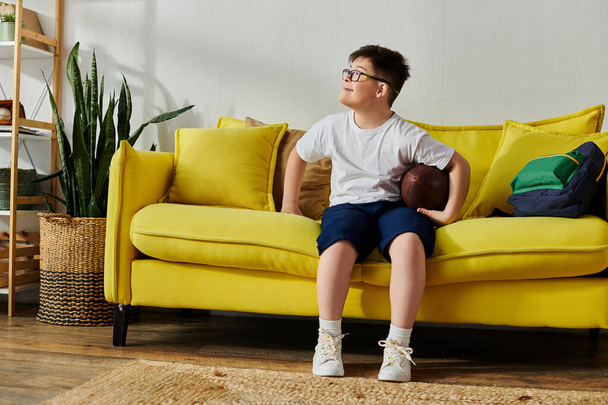 Egy bájos Down-szindrómás fiú, aki egy focilabdát tart, miközben egy sárga kanapén ül.. - Fotó, kép