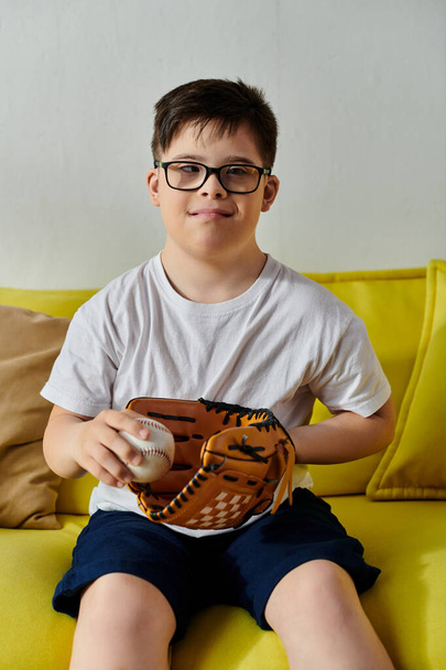 野球を保持している黄色いソファに座っているメガネとダウン症候群を持つ愛らしい少年. - 写真・画像