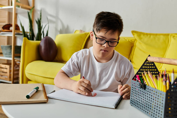 Мальчик с синдромом Дауна сидит за столом, сосредоточенный на записи в блокноте. - Фото, изображение