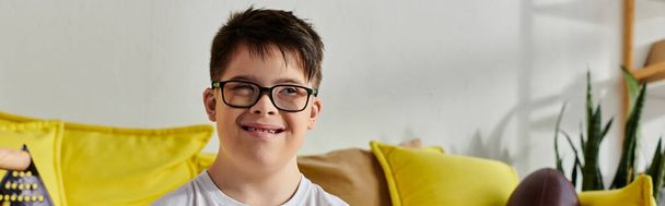 Очаровательный мальчик с синдромом Дауна в очках спокойно сидит на диване в уютной комнате. - Фото, изображение