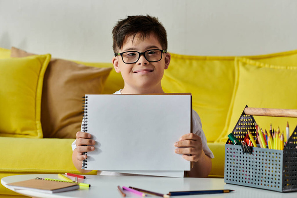 Un adorabile ragazzo con la sindrome di Down mostra orgogliosamente un foglio di carta davanti a un divano giallo. - Foto, immagini