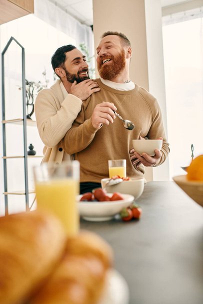 Dos hombres se abrazan en la cocina mientras disfrutan del desayuno juntos en un apartamento moderno, expresando su amor y afecto. - Foto, imagen