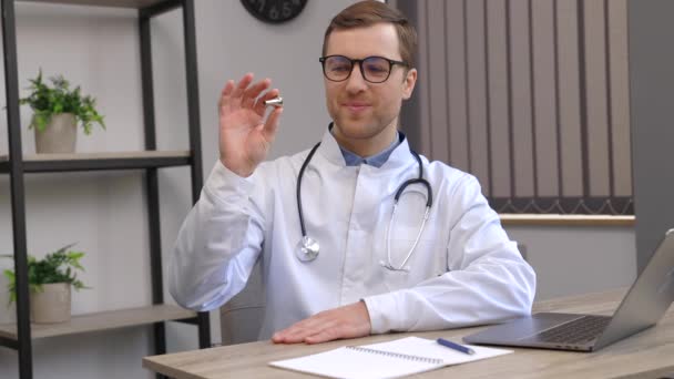 Νεαρός ελκυστικός γιατρός otolaryngologist κάθεται στο χώρο εργασίας του στο γραφείο και κρατώντας ένα ακουστικό χωνί στα χέρια του - Πλάνα, βίντεο