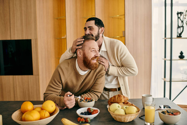 Δύο άντρες, ένα ευτυχισμένο ζευγάρι γκέι, κάθονται σε ένα τραπέζι σε ένα μοντέρνο διαμέρισμα, απολαμβάνοντας το πρωινό μαζί.. - Φωτογραφία, εικόνα