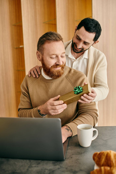 Dois homens trocam um presente na frente de um laptop em um apartamento moderno, compartilhando um momento de felicidade e amor. - Foto, Imagem