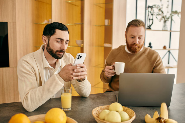 Szczęśliwa para gejów siedzi przy stole w nowoczesnym mieszkaniu, pochłonięta telefonami, ciesząc się dobrym czasem spędzonym razem. - Zdjęcie, obraz