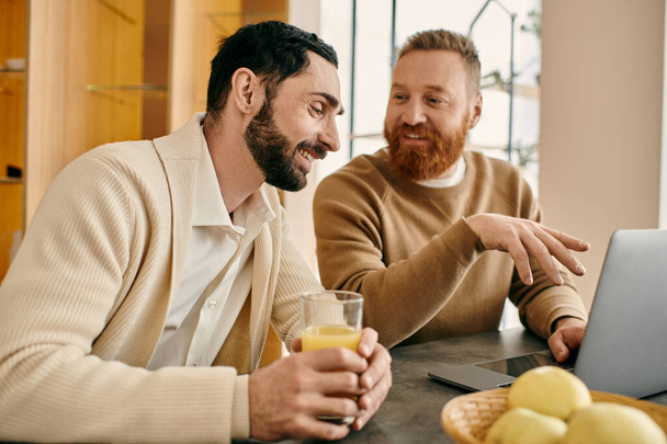 Dva muži šťastně angažovaní, sedící u stolu, zaujatí na obrazovce svého notebooku v moderním bytovém prostředí. - Fotografie, Obrázek