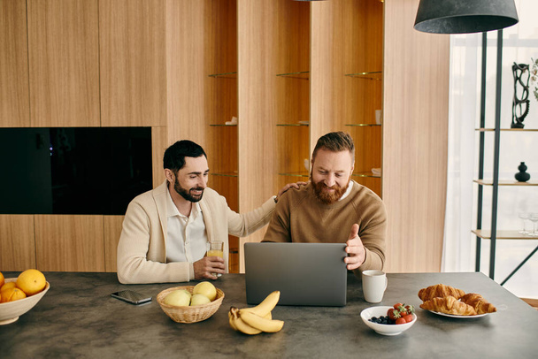 Dwóch szczęśliwych mężczyzn, para gejów, siedzi przy stole w nowoczesnym mieszkaniu, pochłonięci korzystaniem z laptopa. - Zdjęcie, obraz