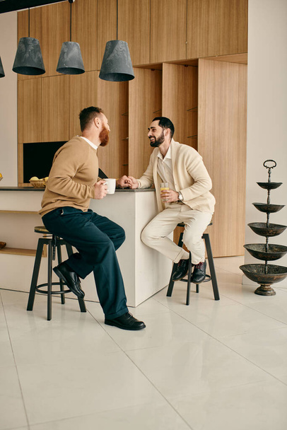 A happy gay couple sitting on stools in a modern kitchen, enjoying each others company and spending quality time together. - Zdjęcie, obraz