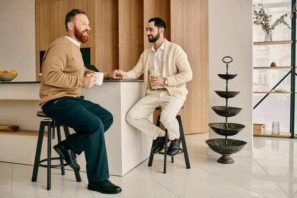 İki adam, mutlu bir eşcinsel çift, modern bir mutfaktaki taburelerde otururken sohbet ediyorlar.. - Fotoğraf, Görsel